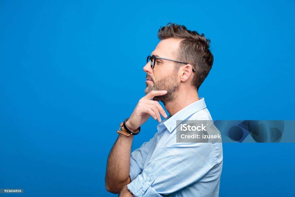 Side view of confident, handsome man, blue background Summer portrait of confident, handsome man wearing blue shirt and glasses, looking away with hand on chin. Side view. Studio shot, blue background. Contemplation Stock Photo