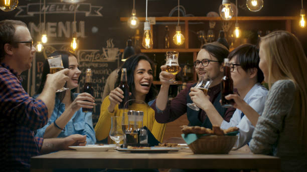 zróżnicowana grupa przyjaciół świętować z toast i clink podniesione okulary z różnych napojów w uroczystości. piękni młodzi bawią się w stylowym barze/ restauracji. - clink zdjęcia i obrazy z banku zdjęć