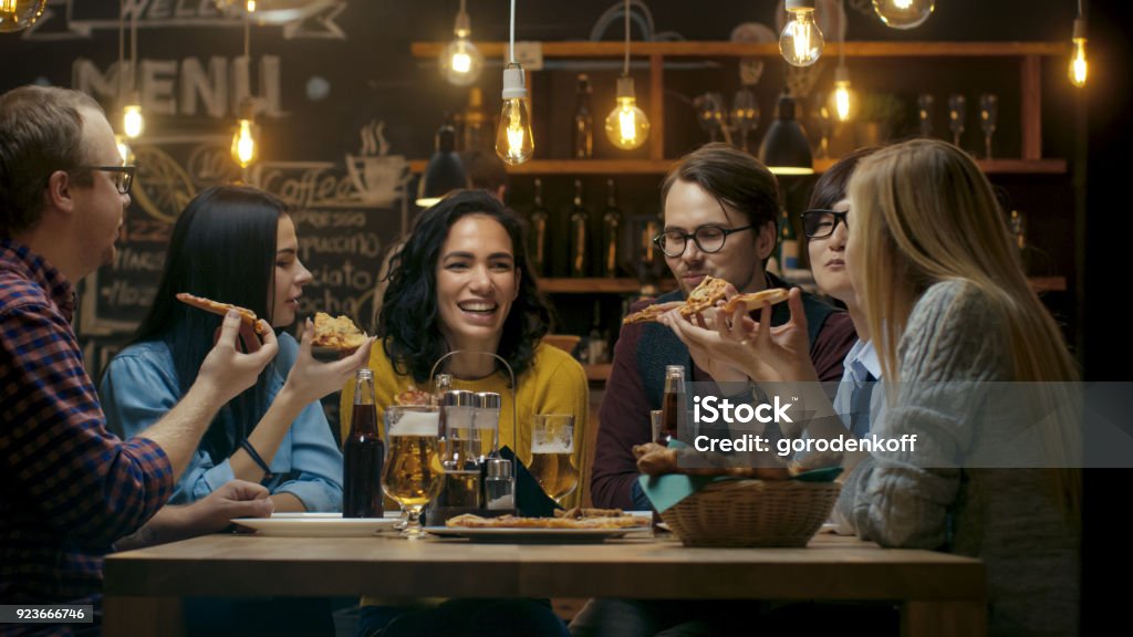 In the Bar/ Restaurant Group of Diverse Young People Eat Slices of Pizza Pie. They Talk, Tell Jokes and Have Fun in This Stylish Establishment. Restaurant Stock Photo