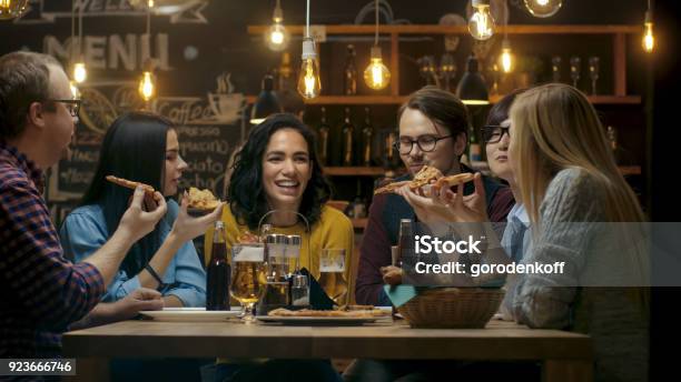 In The Bar Restaurant Group Of Diverse Young People Eat Slices Of Pizza Pie They Talk Tell Jokes And Have Fun In This Stylish Establishment Stock Photo - Download Image Now