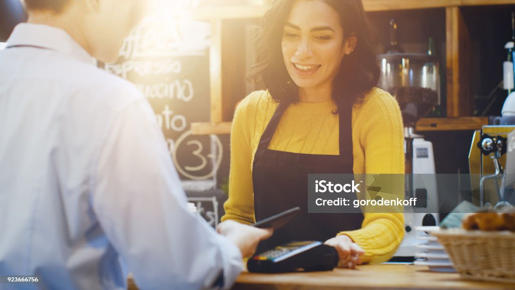 In the Cafe Beautiful Hispanic Woman Makes Takeaway Coffee For a Customer Who Pays by Contactless Mobile Phone to Credit Card System. Store Stock Photo