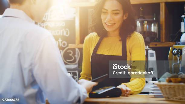 Foto de No Café Bela Mulher Hispânica Faz Café Para Levar Para Um Cliente Que Paga Pelo Celular Sem Contacto Para Sistema De Cartão De Crédito e mais fotos de stock de Loja