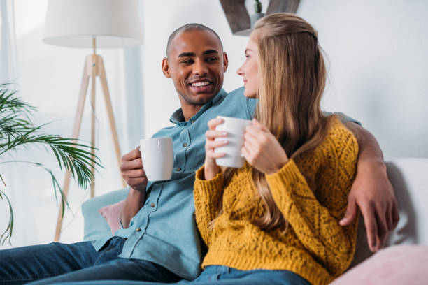 istock multicultural couple sitting on sofa with coffee and looking at each other 923677710