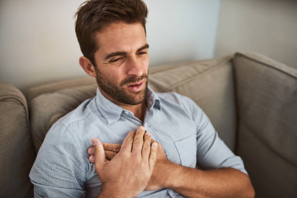 Chest pain is never good Shot of an uncomfortable looking young man holding his chest in discomfort while being seated on a couch at home never stock pictures, royalty-free photos & images