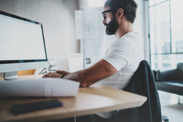 istock Young bearded professional architect wearing eye glasses working at modern loft studio-office with desktop computer.White blank display screen.Blurred background. Horizontal. 923693548