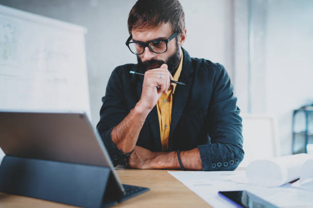 istock Pensive bearded male architect wearing eye glasses working on a digital tablet dock at his desk. Professional experienced engineer constructionist developer.Horizontal.Blurred background. 923698230