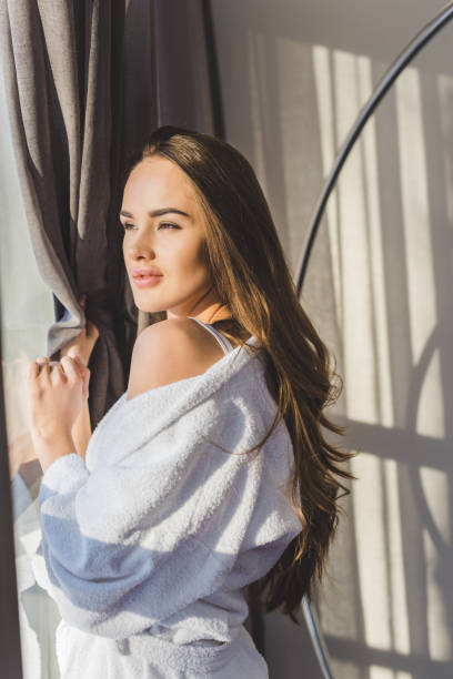 istock pretty pensive woman in bathrobe standing at window at home 923705162