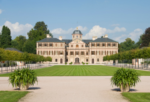 Schwetzingen, Germany - October 13, 2021: Schwetzingen Castle Park. Orangery, built by Karl Theodor. The walls are decorated with facade paintings and simulate natural stone. In the foreground a green meadow with statues framed by a small creek.