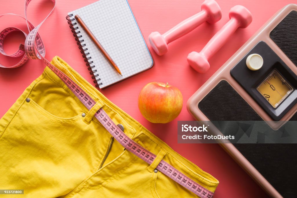 Measure tape with yellow jeans, dumbbells, weight scale, apple, notebook on a pink background. Women diet before summer season. Healthy lifestyle, body slimming, weight loss concept. Cares about body. Body Mass Index Stock Photo