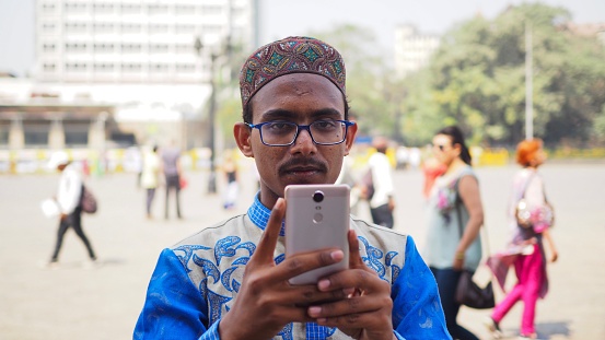 Indian local man wearing traditional cloth holding a smartphone to take photography of the photographer. The scene was at gate of India in Mumbai on the 21st February 2018.