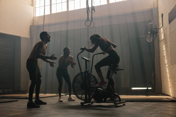 friends motivating woman on exercise bike in gym - spinning instructor exercising gym imagens e fotografias de stock