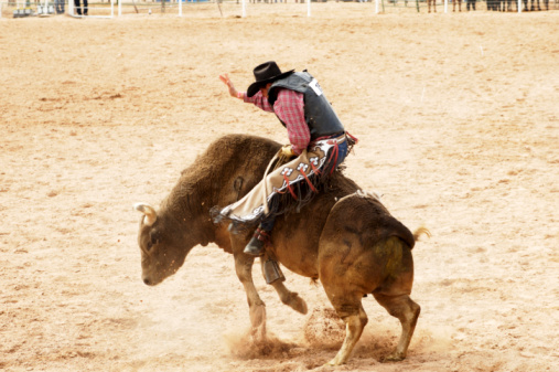 A man in a western attire riding a bull in a rodeo stadium with people spectating in the background.