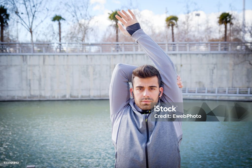 Young athlete man is doing sport runing One young caucasian athlete man is warming up near the river before running in a park Active Lifestyle Stock Photo