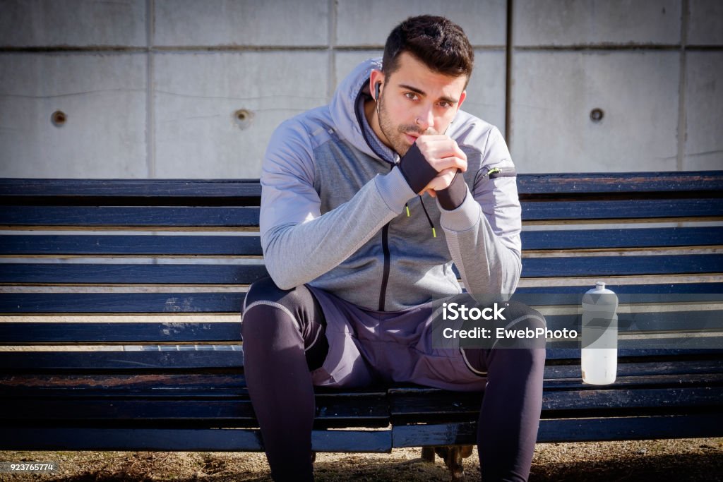 Young athlete man is doing sport runing One young caucasian athlete man is resting on a bench with a water bottle after running in a park Active Lifestyle Stock Photo