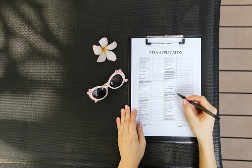 bright flat lay of woman hands fill visa application with pen for travel, sunlight portrait with sunglasses and flower. concept of traveling journey. Front used with Open Font License