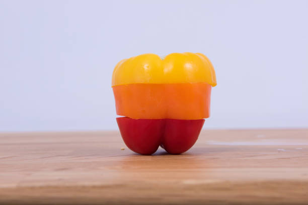 Stock photo showing close-up view of a heap of whole, shiny green, orange and red bell peppers (capsicums), with stems on a red surface, pictured against a mottled blue background. Peppers are a particularly versatile vegetable, often being eaten raw or cooked. Healthy eating concept.