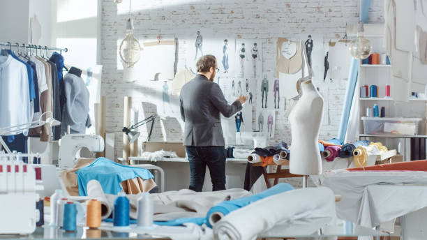 moda hombre, diseñador, bocetos de ropa pernos a la pared. su estudio es sunny, ordenador personal muestra su obra. coloridos tejidos, ropa colgando y artículos de costura son visibles. - garment mannequin men store fotografías e imágenes de stock