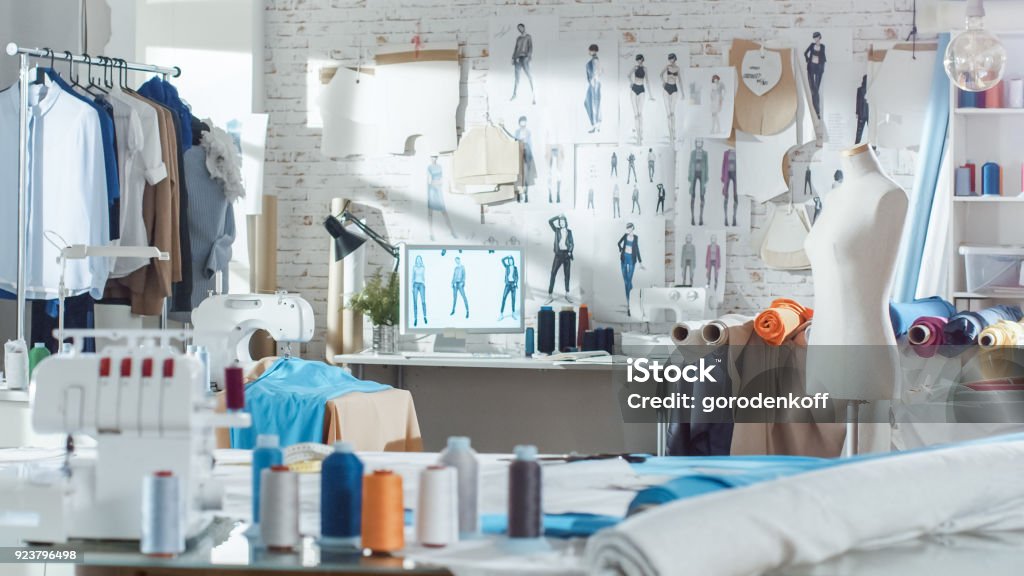 Shot of a Sunny Fashion Design Studio. We See Working Personal Computer, Hanging Clothes, Sewing Machine and Various Sewing Related Items on the Table, Mannequins Standing, Colorful Fabrics. Fashion Stock Photo