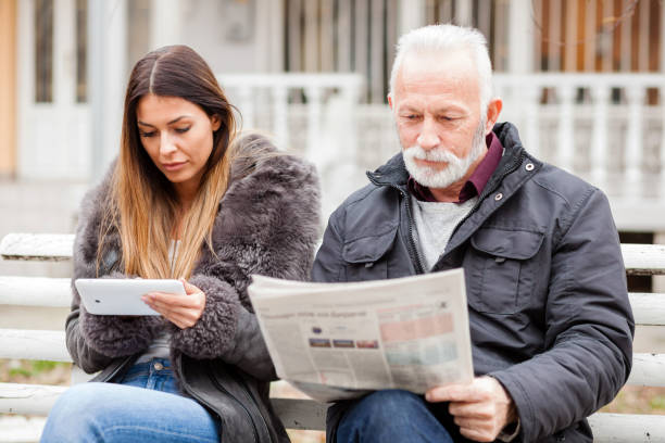 istock Young woman and senior man sitting on bench, reading newspapers and using a tablet 923803400