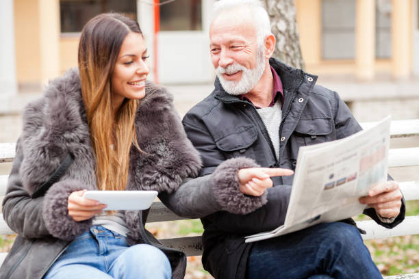 istock Young woman and senior man sitting on bench, reading newspapers and using a tablet 923803534
