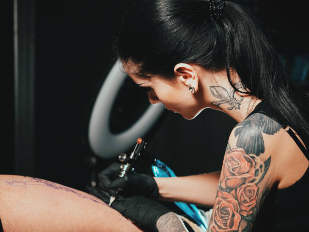 cerca artista del tatuaje muestra el proceso de tatuaje negro con pintura. concentrado contorno femenino para colorear con tinta negra en la pierna con pistola - tattoo gun fotografías e imágenes de stock