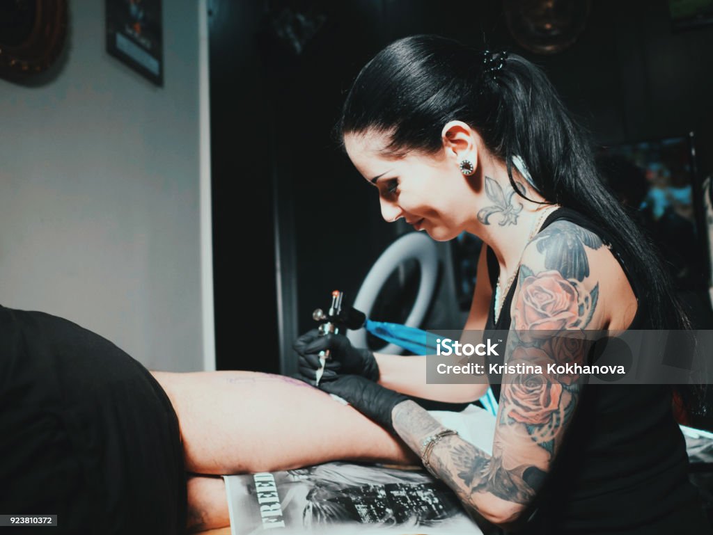 Close up of artist demonstrates the process of getting black tattoo with paint. Master working in workshop. Tattooing young man on leg. Concept: studio, customer communication, freedom, art, young Tattooing Stock Photo