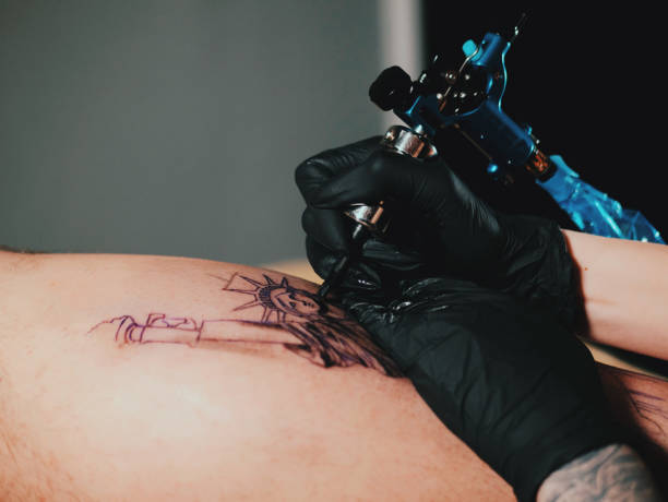 el artista del tatuaje en el trabajo. mujer en guante de látex negro tatuaje estatua de la libertad a una joven hermosa en estudio. cierre para arriba - statue liberty audio fotografías e imágenes de stock