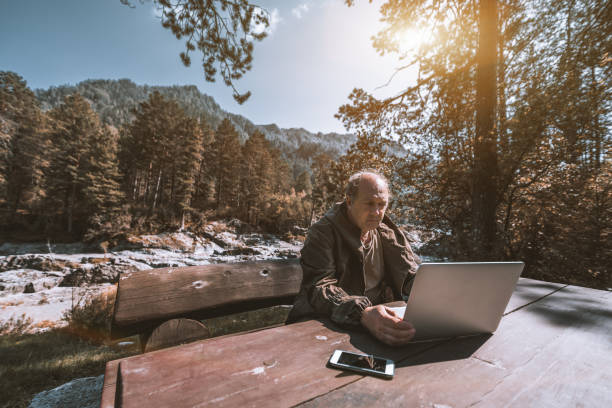 Old hunter with his laptop in mountains Wide-angle view of mature forester sitting alone at wooden table and working with his laptop on sunny autumn day; Caucasian aged hunter in forest near mountain river with his netbook and smartphone woodward stock pictures, royalty-free photos & images