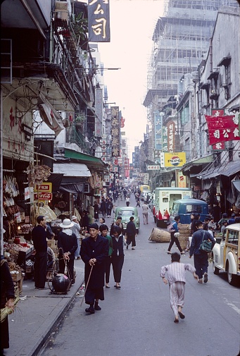 Hong Kong, China, 1966. Street scene in Hong Kong with pedestrians, shops, advertising signs, buildings, car, dealers and goods.