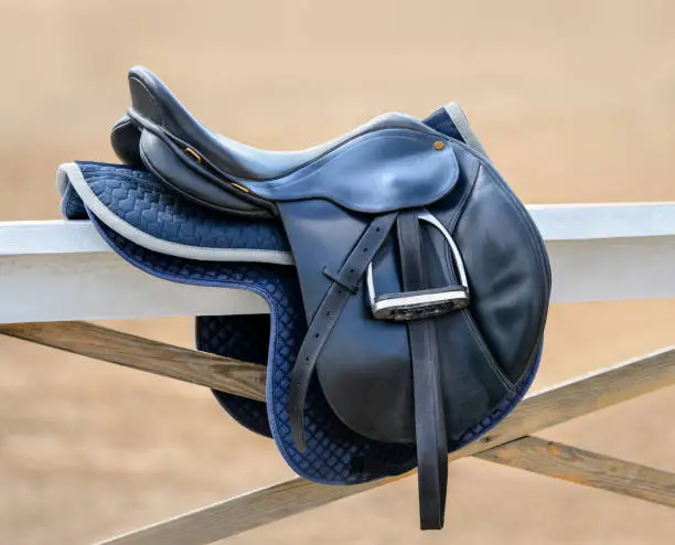 Black English saddle hanging on fence near stables. Side view.