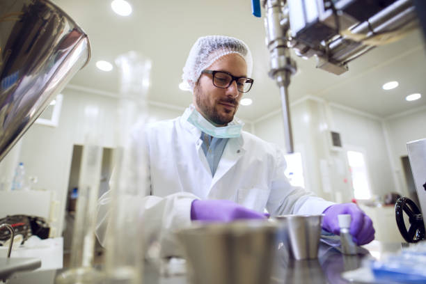 feche de jovem cientista profissional sério focado barbuda sentado e trabalhando na máquina do laboratório com metais frascos e garrafas na frente. - máquina de manufactura farmacêutica - fotografias e filmes do acervo