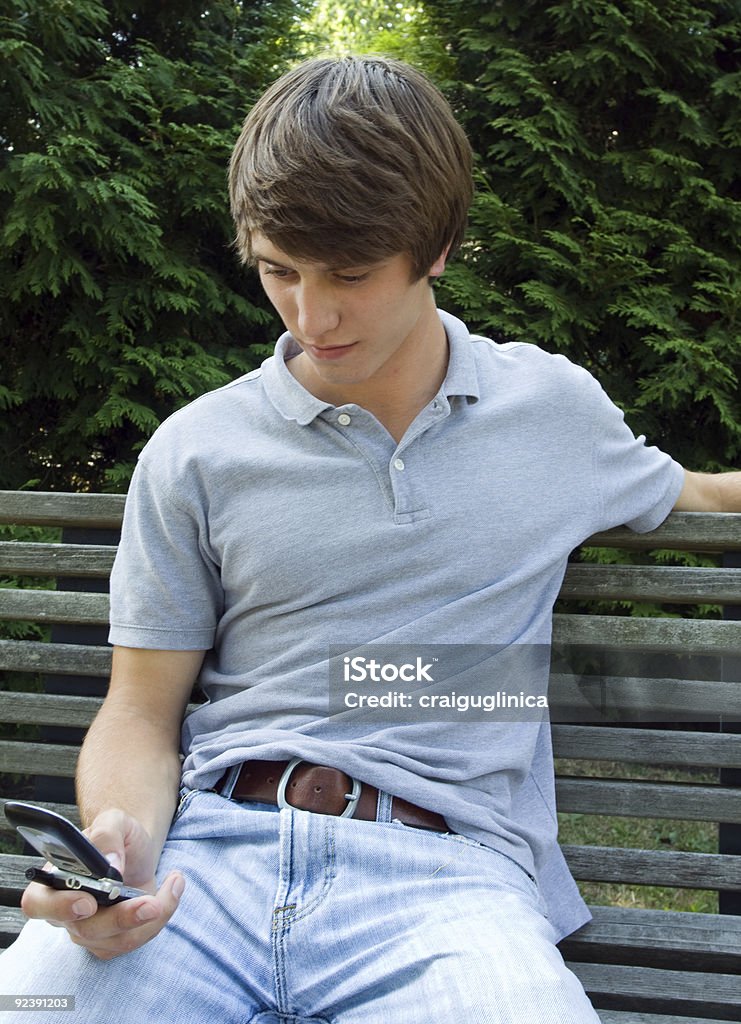 Teen text Handsome young male texting or dialing cell phone while sitting alone on a park bench in the summer. Adults Only Stock Photo