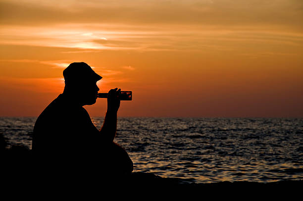 Close-up of hand holding beer bottle against sky. Cropped image of mid adult man is enjoying alcohol in yard. Focus is on limb during sunny day. Man has tattoo in his forearm.