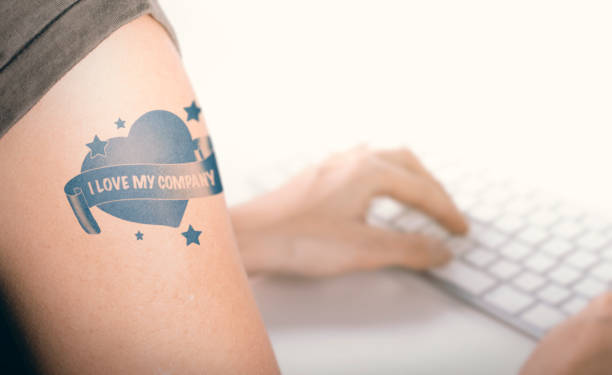 Employee Loyalty and Engagement Woman working on a computer with close up on a tatoo with the text i love my company.  Concept of employee engagement and feeling good at work retain stock pictures, royalty-free photos & images