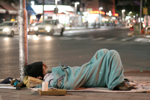 Homeless Male With Sign Sleeping Near Garbage And Rags