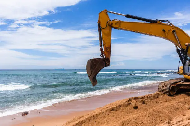 Construction excavator machine on beach water line coastline building repairs with sandbags buffer on sand dunes to protect road from ocean storms.