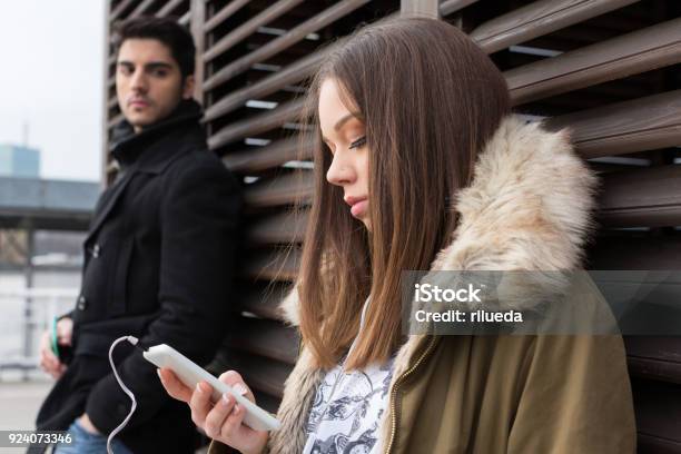 Young Woman Looking At Mobile Phone Man Jealous In The Background Stock Photo - Download Image Now