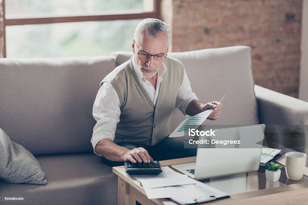 Old confident clever concentrated bearded grandpa is checking his calculations sitting in front of monitor at office Senior Adult Stock Photo