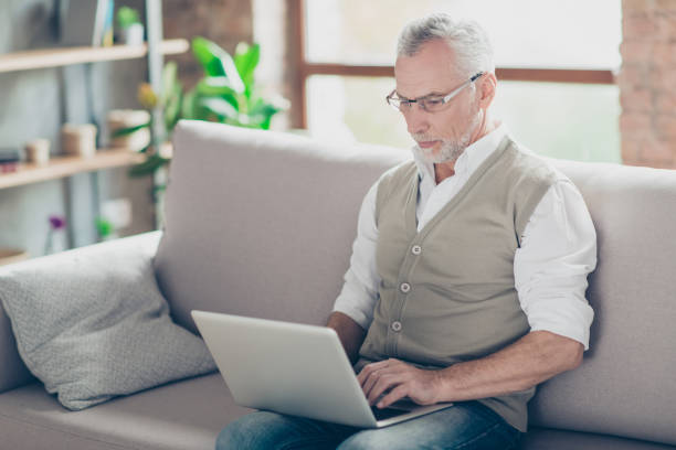 Half-turned photo of experienced qualified clever serious pondering thinking analyzing confident planning granddad holding netbook on knees sending getting receiving emails sitting on sofa