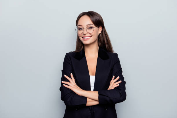 Portrait of young, caucasian, stunning, nice stylish, sexy headmistress in black jacket, formal wear with crossed arms and beaming smile standing over grey background stock photo