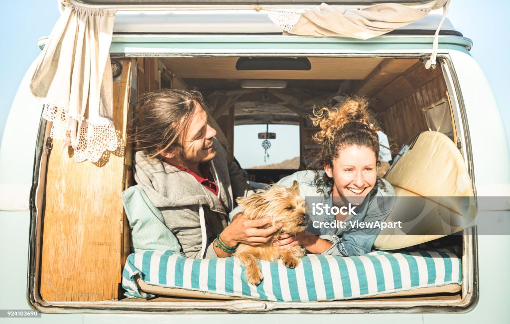 Hipster couple with cute dog traveling together on vintage mini van transport - Life inspiration concept with hippie people on minivan adventure trip in relax moment - Bright warm retro filter Camping Stock Photo