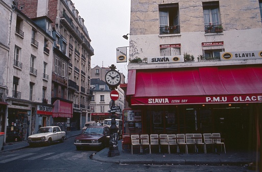 Paris, France, 1982. Street scene with clock, bistro, buildings, cars, pedestrians, shops and advertising in the heart of Paris.