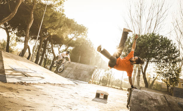 urban athlete breakdancer performing acrobatic jump flip at skate park - guy riding bmx bicycle behind mate acrobat dancing with extreme move - breakdance and friendship concept - warm sunset filter - bmx cycling bicycle street jumping imagens e fotografias de stock