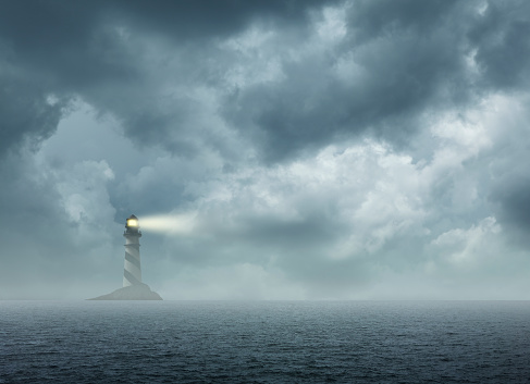 A lighthouse sits on a small rocky island on the horizon as it shines its light during inclement weather.