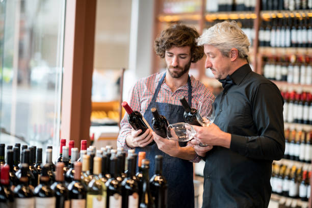 young sales clerk and business owner looking at bottles of wine while owner holds wine glasses at a winery - sales clerk imagens e fotografias de stock