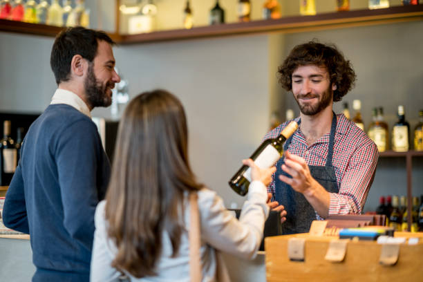 Couple at a wine store checking out purchasing a bottle of white wine and friendly salesman helping them Couple at a wine store checking out purchasing a bottle of white wine and friendly salesman helping them all looking very happy and smiling liquor store counter photos stock pictures, royalty-free photos & images