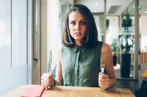 Hungry young woman having lunch in restaurant, holding knife and fork. Looking at camera.