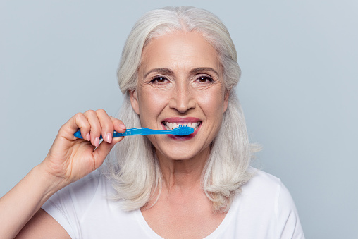 Concept of cleaning teeth is a correct way. Close up photo of happy cheerful mature old lady with good visage brushing her teeth with a blue toothbrush, isolated on gray background