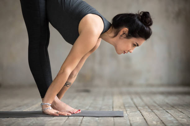 Young woman practicing yoga, doing Ardha uttanasana exercise, Standing Half forward pose, working out, wearing sportswear, black pants and top, indoor close up photo, gray wall in yoga studio