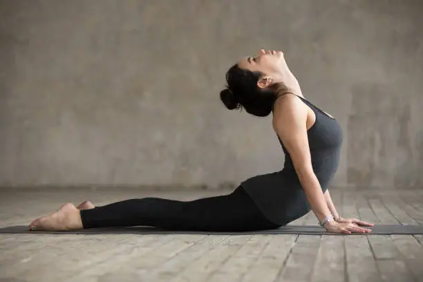 Young woman practicing yoga, doing Cobra exercise, Bhujangasana pose, working out, wearing sportswear, black pants and top, indoor full length, gray wall in yoga studio